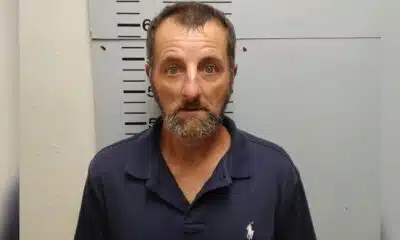 A man with a beard and blue polo shirt stands against a height chart in a police booking photo. — Shooting girlfriend