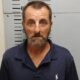 A man with a beard and blue polo shirt stands against a height chart in a police booking photo. — Shooting girlfriend