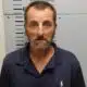 A man with a beard and blue polo shirt stands against a height chart in a police booking photo. — Shooting girlfriend