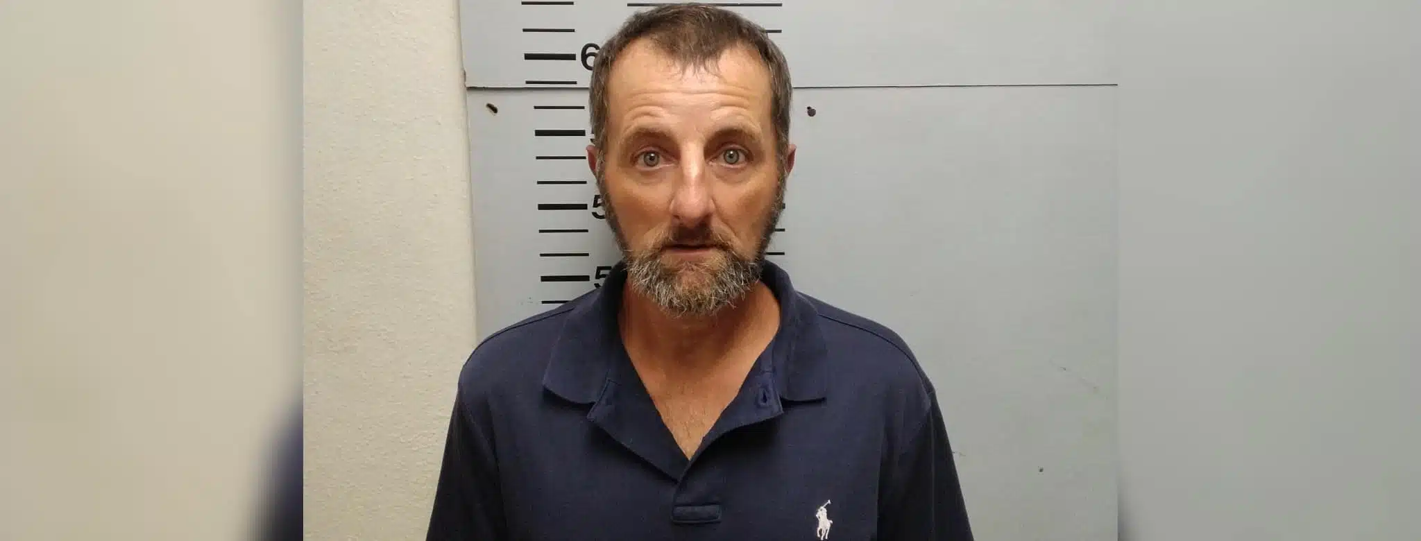 A man with a beard and blue polo shirt stands against a height chart in a police booking photo. — Shooting girlfriend