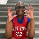 Pharrah Moody in a red Lady Vikes basketball jersey and glasses, smiling and gesturing on gym bleachers
