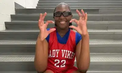 Pharrah Moody in a red Lady Vikes basketball jersey and glasses, smiling and gesturing on gym bleachers