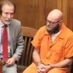 Joey Foster in an orange jail uniform sits beside his attorney in a wood-paneled courtroom.