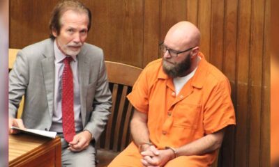 Joey Foster in an orange jail uniform sits beside his attorney in a wood-paneled courtroom.