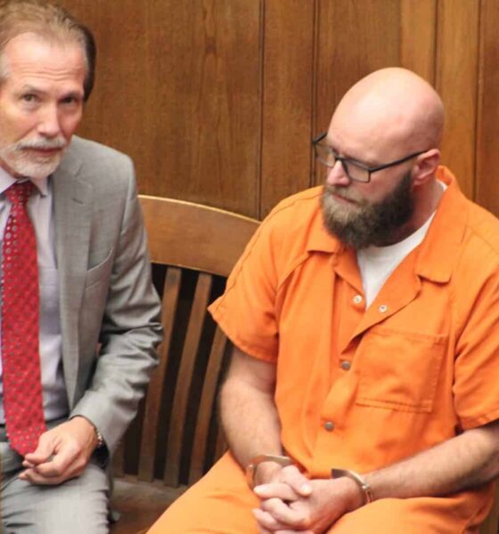 Joey Foster in an orange jail uniform sits beside his attorney in a wood-paneled courtroom.