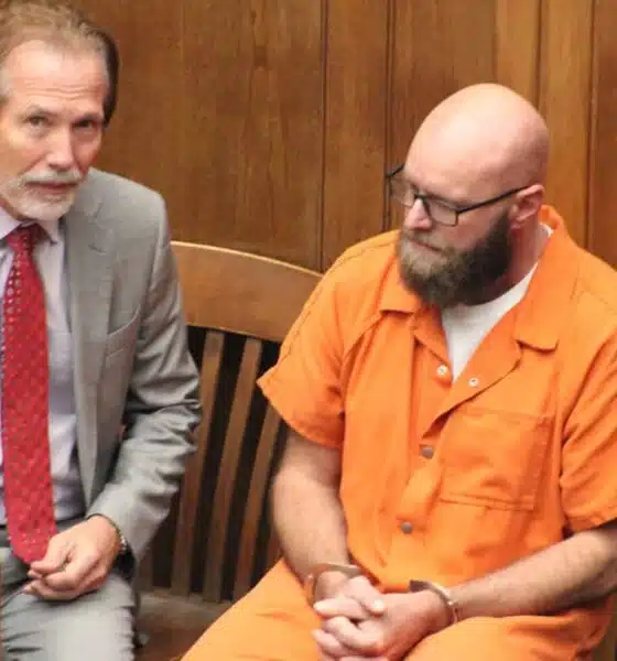 Joey Foster in an orange jail uniform sits beside his attorney in a wood-paneled courtroom.