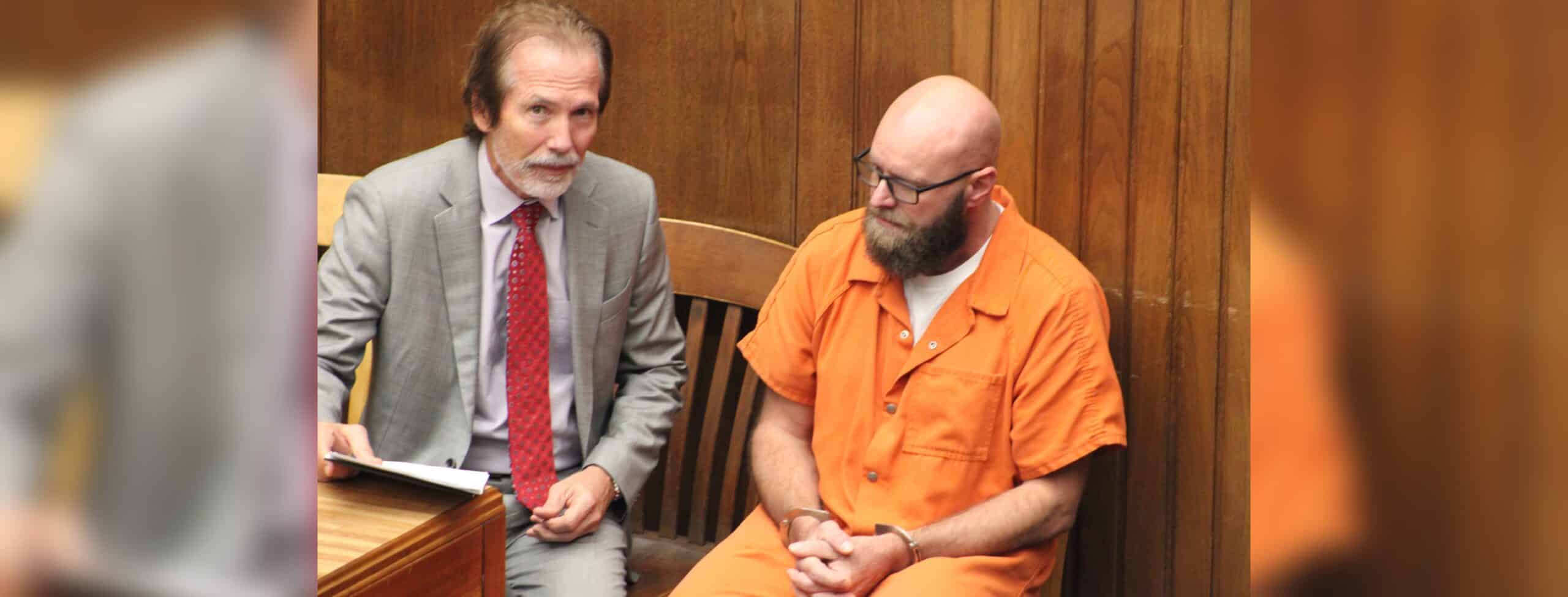 Joey Foster in an orange jail uniform sits beside his attorney in a wood-paneled courtroom.