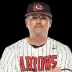 A coach in a striped baseball jersey and cap, representing Hinds Athletics, poses confidently with arms crossed.