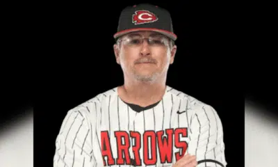 A coach in a striped baseball jersey and cap, representing Hinds Athletics, poses confidently with arms crossed.