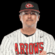 A coach in a striped baseball jersey and cap, representing Hinds Athletics, poses confidently with arms crossed.