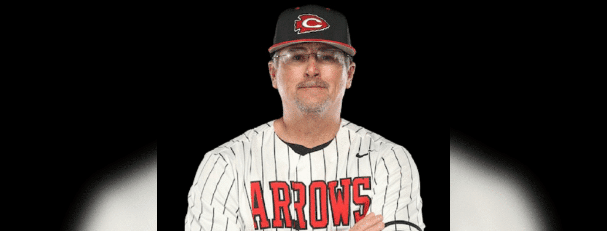 A coach in a striped baseball jersey and cap, representing Hinds Athletics, poses confidently with arms crossed.