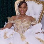 A young woman in an ornate white-and-gold gown sits on a throne-style chair beside black and gold balloons. — Milah Davis