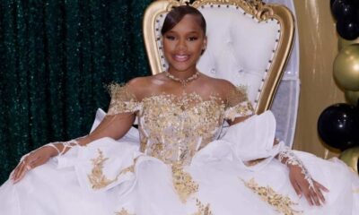 A young woman in an ornate white-and-gold gown sits on a throne-style chair beside black and gold balloons. — Milah Davis