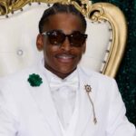 Cameron Pickett in a white suit and sunglasses seated in an ornate chair