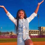 Addison Anderson poses with arms raised on a track in front of a stadium on a sunny day.