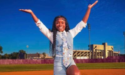 Addison Anderson poses with arms raised on a track in front of a stadium on a sunny day.