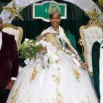 Gatotbration ceremony display with a student in a white gown and crown between two adults, amid black and gold balloons