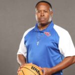 WC freshmen boys basketball coach in a blue-and-white polo holds a basketball against a gray backdrop.