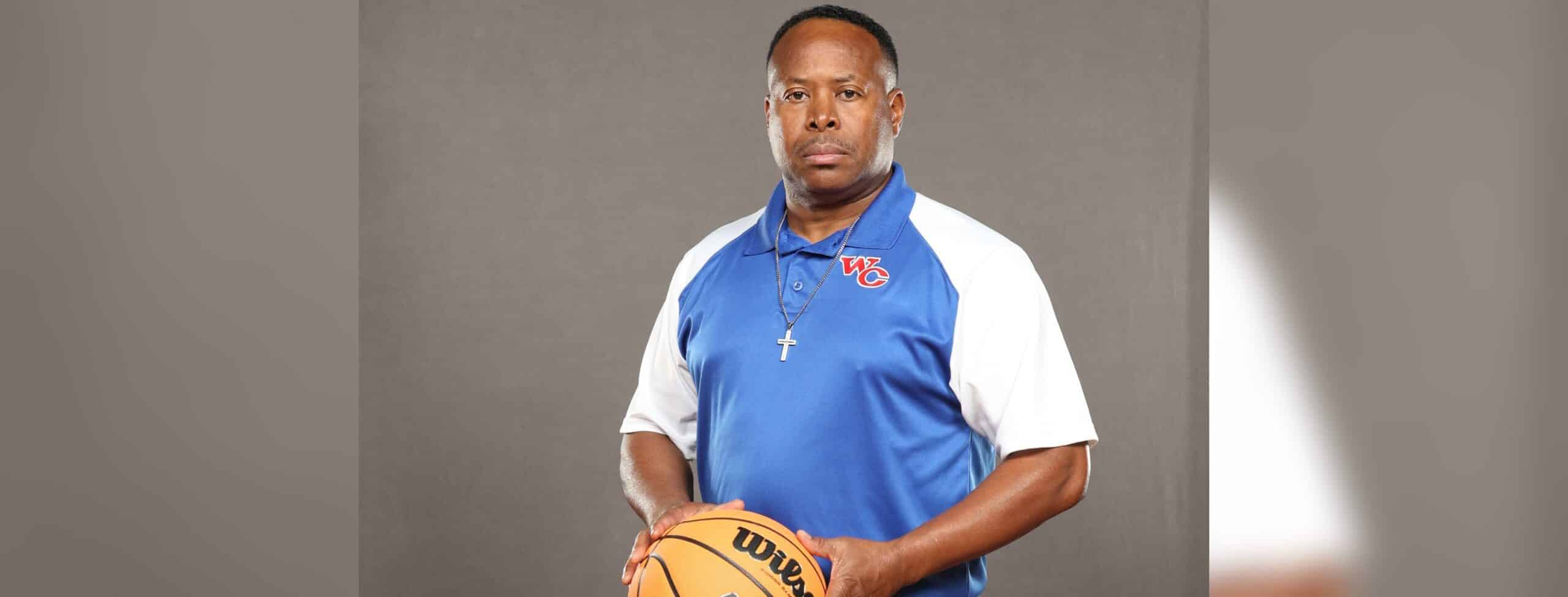 WC freshmen boys basketball coach in a blue-and-white polo holds a basketball against a gray backdrop.