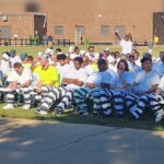 About 300 inmates in striped uniforms and white shirts sit in rows outdoors for a graduation ceremony.