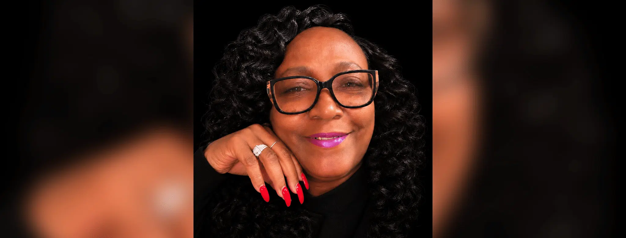 A woman with curly hair and glasses smiles, showcasing her long red nails and a ring. — Stephanie Cherey Jones